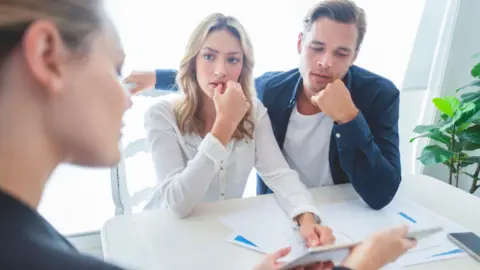 Getty Images Otra mujer muestra a un hombre y una mujer preocupados, de unos 30 años, una tableta con papeles sobre la mesa frente a ellos.