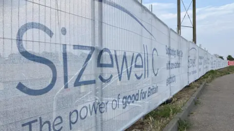 Guy Campbell/BBC A construction fence with white fabric over it blocking off the view behind it. The words "Sizewell C, The power of good for Britain" are written in blue writing on the white fabric.