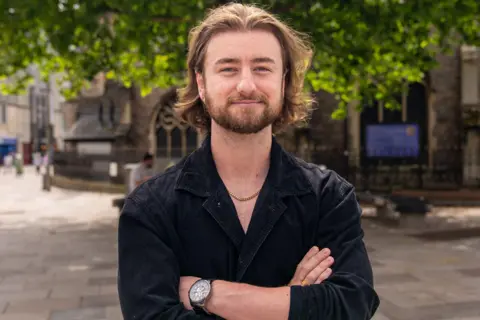 Tom crosses his arms stood in a city centre. He has a watch on his left wrist and a gold chain around his neck. He is wearing a black shirt and has long flowing brown hair.