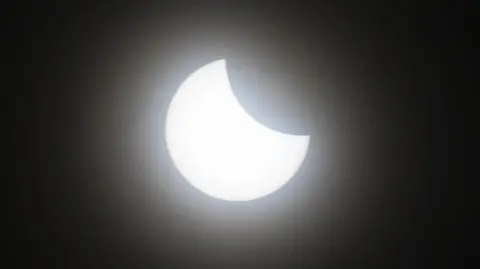 A picture of a partial eclipse showing a bright white sun with a crescent over its upper-left quarter. The background is black. 