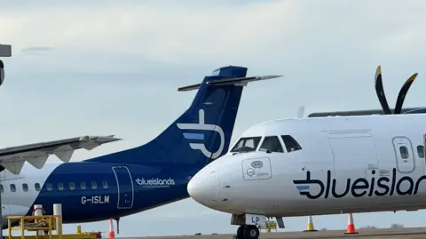 Two aircraft are stationary at Guernsey Airport. One is coloured navy blue with he logo of Blue Islands in white on the tail. The logo is an interlocking letter B and I. The second aircraft is white with the words Blue Islands in navy blue lettering.
