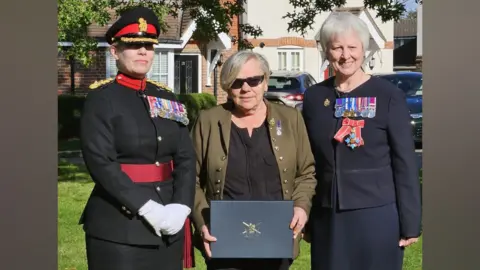 Carol Morgan Three women stood in a line. On the left, a woman is wearing a black and white army uniform. In the centre, the woman is holding a box with a crest on it. On the right, the woman is wearing a line of army medals.
