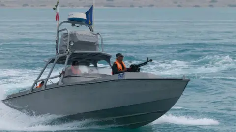 A boat patrols the waters off an arid coast, with a man in a bright orange jacket onboard holding a machine gun