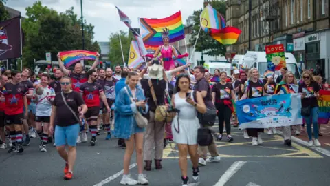 The Newcastle Pride march in 2024. Crowds of people are walking down the middle of the road towards the camera. They are wearing brightly coloured cloths and are waving rainbow flags. 