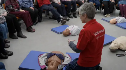 Female volunteer Carol Wood, with grey hair and glasses, has her back turned to the camera. She is kneeling on the floor and is wearing a red polo shirt which says Volunteer on the back. On the floor in front of her is a female manikin to practice CPR on. 