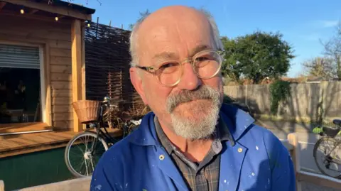 Vikki Irwin/BBC David Riches in a blue overall over a checked brown shirt wears glasses. He has a beard. He is standing in front of a wooden garage with a bicycle behind him.