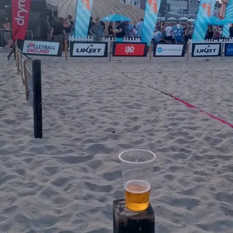 Weymouth and Portland Marine Litter Project A plastic cup placed on a wooden post containing about a third of a pint of beer. In the background is the beach and volleyball arena bearing the names of sponsors around the edge.