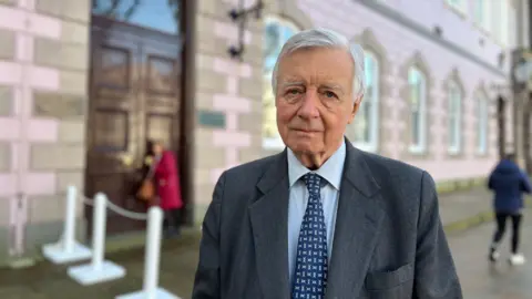 Former Bailiff of Jersey, Sir Philip Bailhache wearing a grey suit and blue shirt and tie. He has short grey hair and is stood in front of the states building in Jersey, which is blurred.