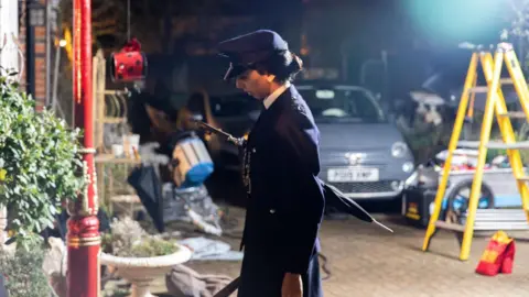 Pinder Productions, Geppetto Pictures, Kai Campbell A women dressing in a old fashioned police officers uniform with an umbrella walking outside on a film set, in the background is a bright spotlight and a ladder propped up.