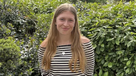 Lillia has long blonde hair down either side and wears an off the sleeve black and white hooped top with a heart pendant necklace. She stands in front of a hedge.