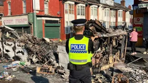 A police man stands looking at a burned down bus