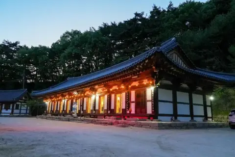 BBC/ Hosu Lee Traditional Korean temple illuminated with warm lights at dusk, set in a quiet courtyard at the edge of a forested hillside