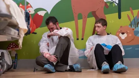 HBO Max Dr Robby (Noah Wyle) breaks down on the floor of the hospital - he is sitting next to Dr Whitaker (Gerran Howell)