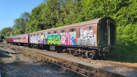 Avon Valley Railway A heritage railway train with three reddish-coloured coaches on a sidling with trees behind. The train has graffiti-style images painted on it, including Margaret Thatcher