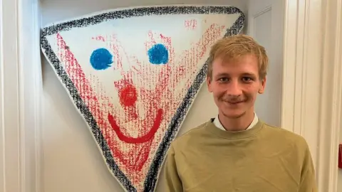 A man in a yellow jumper, with part of a white and blue shirt just showing, is standing in front of a plain background that also includes a drawing of a face in red, white and blue in a triangle.