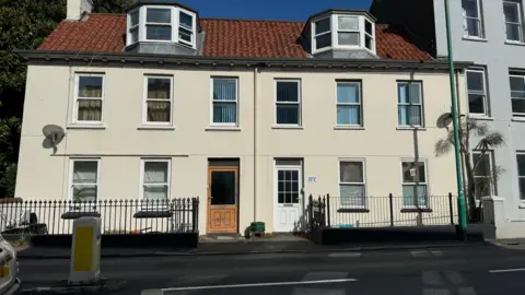 A cream house with 12 windows and two doors, with a black fence in front of it. 