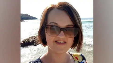 Chloe Morrison, who is wearing sunglasses, takes a selfie on a beach. The sea is in the background.