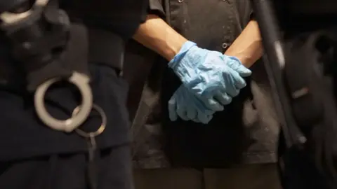 Close up of an immigration officer and the handcuffs hanging off his belt. In the background is someone wearing blue food prep glove and is holding their hands crossed in front of them.