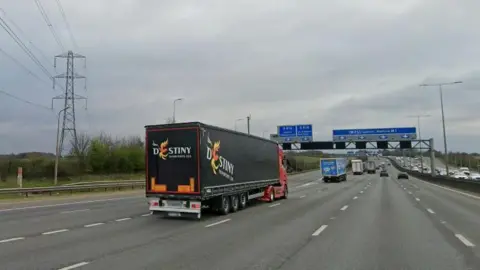 Three lanes of the M1 with two slip roads to the left, and blue motorway signs overhead. There is a line of lorries in the left-hand lane and cars in the other lanes.