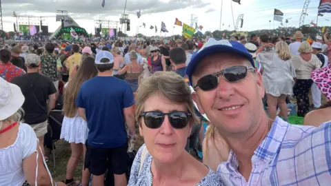 Paddy Gordon Paddy Gordon (right) stands with his wife Maddy in a crowd at Glastonbury. He wears a blue baseball cap, sunglasses and blue, pink and white shirt. She has short light brown hair and is wearing sunglasses and a blue-and-white blouse.