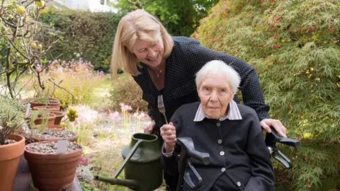 Anita Corbin An elderly woman in a wheelchair holds up a fork. She is in a pretty garden with pots and bushes. 