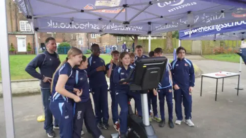 The King's School A group of pupils at The King's School in Gloucester, wearing their blue P.E kit uniform. There is a girl sitting on a static bike underneath a blue cabana, while the other children surround her and cheer her on. 