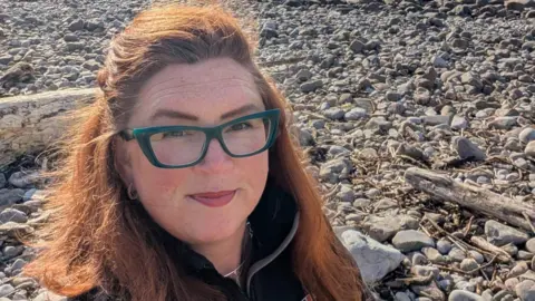 Close up of selfie of Sian smiling whilst stood on a pebbled beach. She has straight ginger hair and wears a black fleece and blue glasses. 