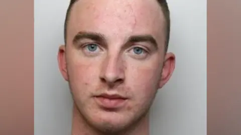 A police mug shot of a young man with short brown hair and blue eyes looking directly into the camera
