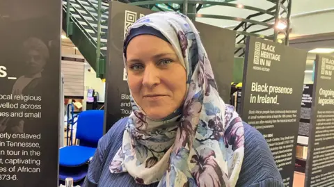 Dr Naomi Green is in a floral headscarf and purple top. 
She is standing beside a number of museum placards. 
A stack of blue chairs and a green staircase are visible behind the signs. 