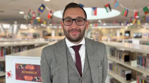 Hayley Clarke/BBC Computer science teacher Sam Alner inside the school library at the Bridge Academy. He is wearing a grey three piece suit and burgandy tie, and smiling at the camera