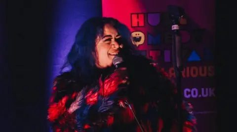 Maxine Wade A young woman with dyed-blue hair wearing a red and black furry jacket talking into a microphone on stage.