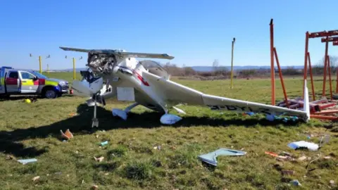 Air Accidents Investigation Branch A crashed light aeroplane on an airfield, surrounded by debris, with bright orange technical structures beside it. There is an emergency vehicle behind the plane and the nose is completely destroyed. It is a sunny day with a blue sky.