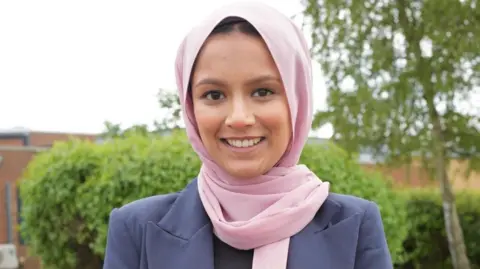 Luton Borough Council Umme Ali with short dark hair wearing a pink headscarf and purple jacket. She is smiling at the camera and standing in front of a green bush and small tree.