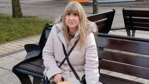 Linda Smith Linda with long blonde hair sat on a bench outside. She is wearing a white coat and can be seen holding a dog lead.