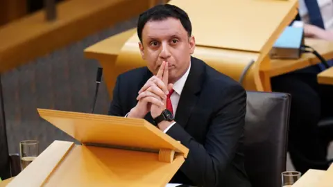 Anas Sarwar sitting in the Scottish Parliament chamber, with his fingers covering his mouth. 