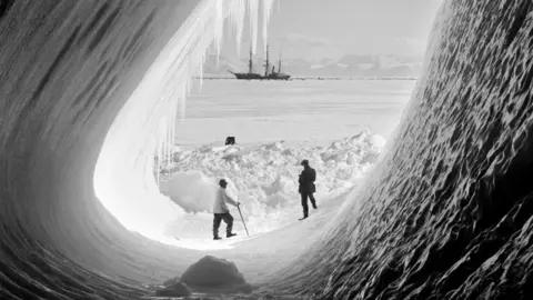 Getty Images/SPRI Black and white image. Two men stand in a huge ice cave. In the background the ice covered sea can be seen with the Terra Nova ship appearing small in the distance  