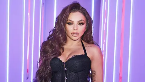 Jesy Nelson is wearing a black top, has long reddish brown hair and is looking at the camera with lip fillers. Behind her is a brightly purple room.