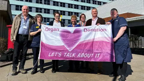 A group of two men and five women, some in uniform, holding a pink and white flag and smiling. They are standing outside a hospital. The flag has the messages "Organ Donation" and "Let's talk about it" on it, and there is a pink heart in the middle.