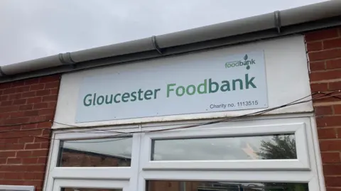 A white plastic sign with Gloucester Foodbank in green letters is stuck to the brick wall above the entrance door.