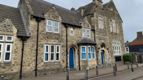 A view of the front of King William Street Church of England Primary School in Swindon. It is a beige-coloured stone building with a blue front door.
