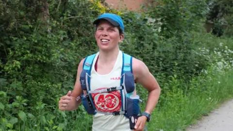 Supplied Laura is running down a gravel track and smiling at the camera. She is wearing a vest top with a red logo on that says 'the triathlon project' along with a blue running vest and cap. 