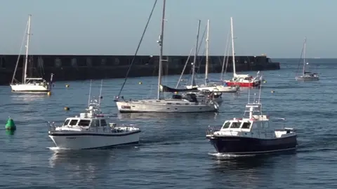 Sorcerer II and Causeway Explorer are sailing side by side into the harbour on a bright sunny day. There are five yacht moored inside the harbour wall.
