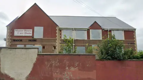 A street view of Manor House which is a large, red brick building with boarded up windows. A Manor House Care Home sign is on the front of the building.