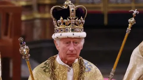 Within the Abbey, King Charles, wearing a bejewelled crown and golden robe, holds two golden sceptres in his hands