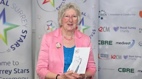 An elderly woman with short white hair, a pink blazer, a blue shirt and clear glasses holding a glass award.