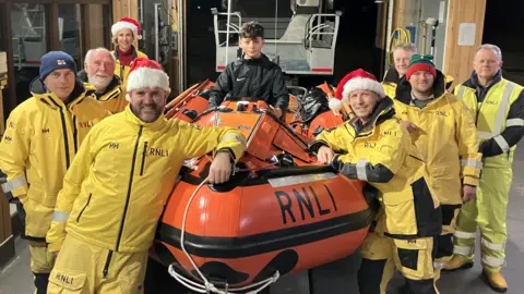 Alex Dunlop/BBC Kaan, a boy who is sitting inside an orange RNLI lifeboat. He is surrounded by a group of eight RNLI volunteers. Some members of the groups are wearing santa hats. 