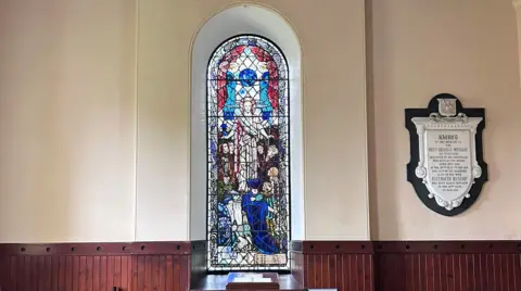 Jackie Wells A more distant view of a stained-glass window in a rural south of Scotland church featuring Jesus at its centre and people gathered in reverence around