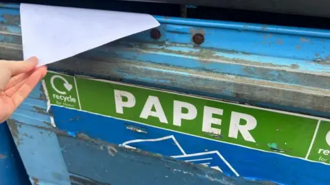 A blue bin with a green sign that reads paper recycle, a hand puts paper in the bin.