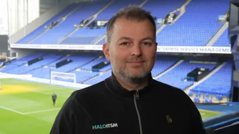 Paul Hamilton standing in a box at Ipswich Town FC ground. He has short greying hair and short greying beard and is wearing a black zipped up top with HaloITSM written on the left side. Beyond him are rows of blue seats and the football pitch.