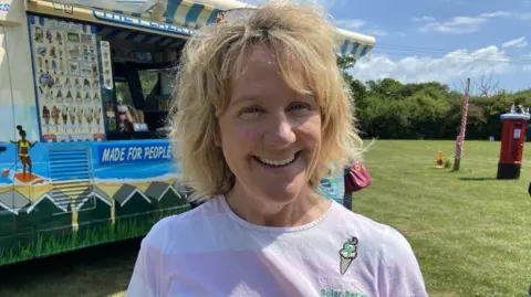 A blonde woman is smiling. She is wearing a light purple tshirt with an icecream logo. There is a red postbox in the background and the sun is shining.